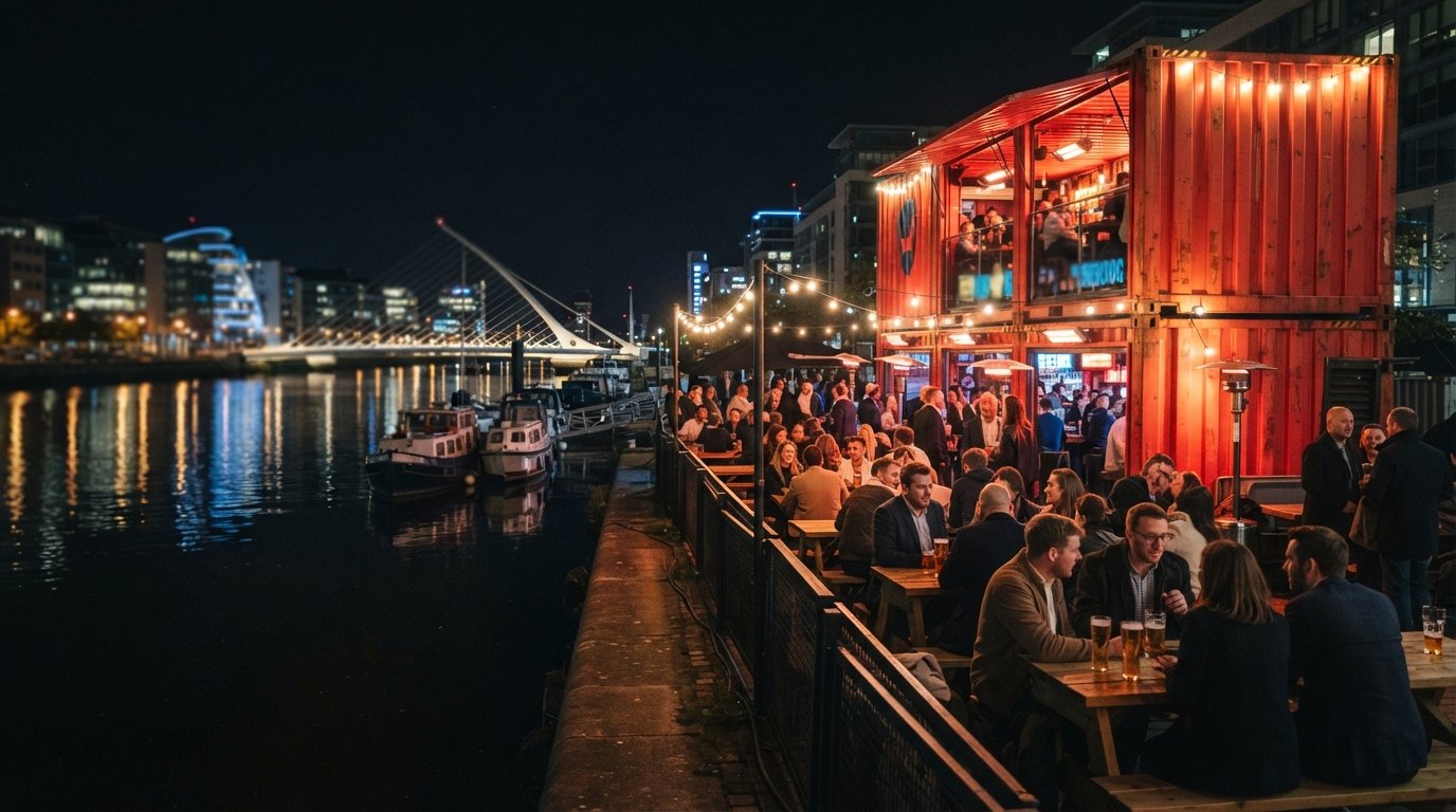 BrewDog Dublin at Capital Dock at night, red shipping-container bar glowing beside the river with the heated terrace and city reflections on the water